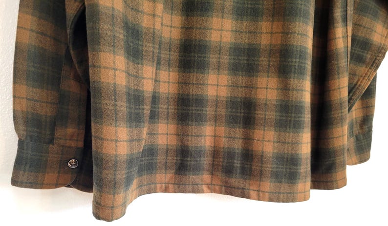 Pode incluir: Close-up de uma camisa de flanela xadrez marrom e verde. A camisa apresenta um padr&atilde;o xadrez cl&aacute;ssico com punho abotoado e um detalhe de bot&atilde;o &uacute;nico. O tecido parece macio e texturizado.