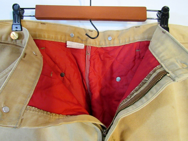 May include: A pair of tan work pants with a red lining and a zipper closure. The pants have multiple pockets and a button fly.