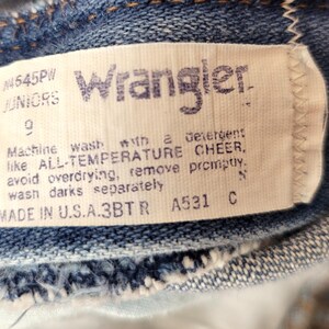 May include: Close-up of a Wrangler jeans tag. The tag is white with blue text, displaying the brand name, size, and care instructions. The jeans are a classic blue denim.