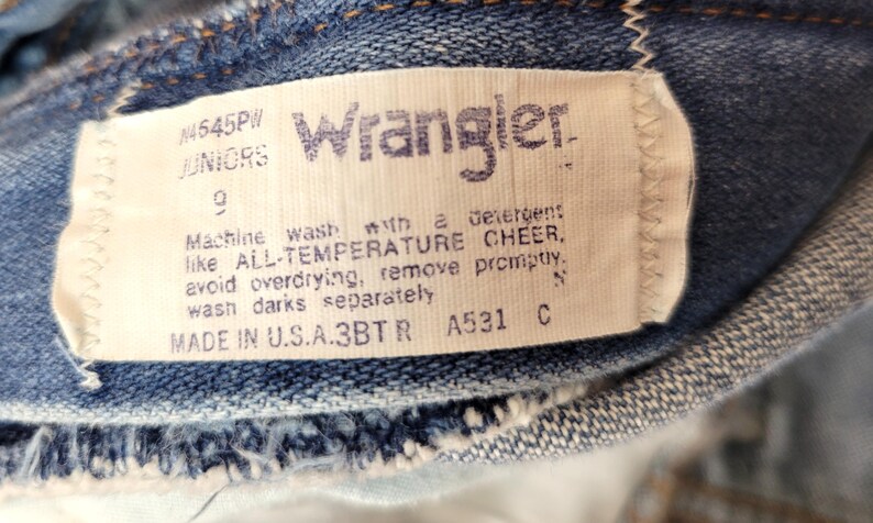 May include: Close-up of a Wrangler jeans tag. The tag is white with blue text, displaying the brand name, size, and care instructions. The jeans are a classic blue denim.