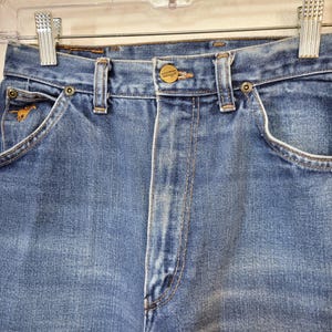 May include: Close-up of light blue denim jeans with a button closure and a small gold-colored horse emblem on the front pocket. The jeans have a classic five-pocket design and are hanging on a clear hanger.