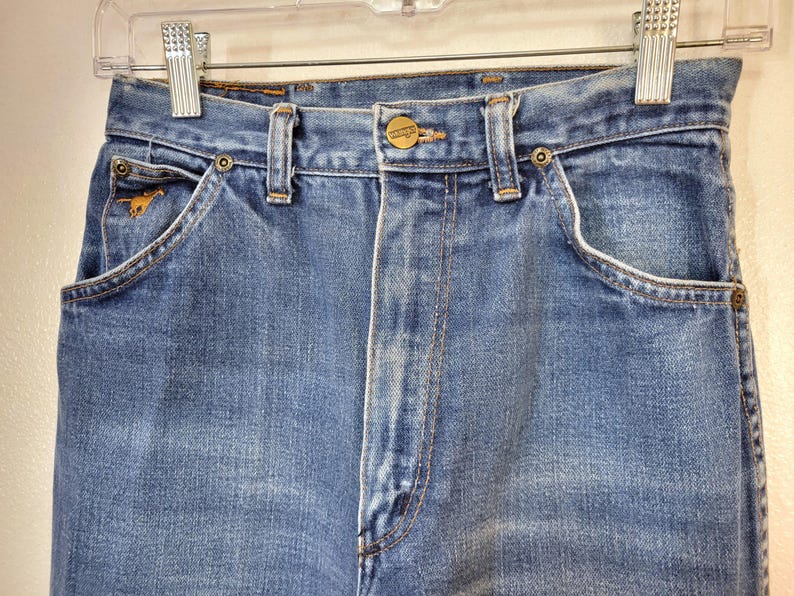 May include: Close-up of light blue denim jeans with a button closure and a small gold-colored horse emblem on the front pocket. The jeans have a classic five-pocket design and are hanging on a clear hanger.