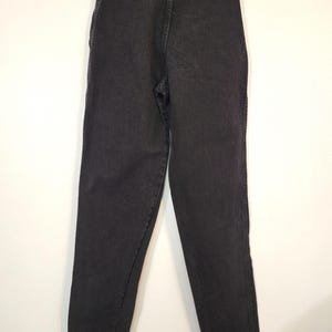 Puede incluir: Un par de vaqueros de mezclilla negros, exhibidos en una percha. Los vaqueros tienen una cintura alta, un dise&ntilde;o cl&aacute;sico de cinco bolsillos y una pernera recta. El logotipo de Wrangler es visible en el bolsillo trasero.