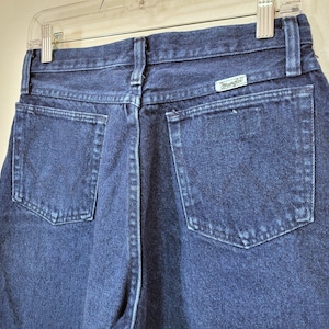 May include: Close-up of the back of dark blue denim jeans. The jeans have two back pockets and a small rectangular tag with the brand name "Wrangler". The jeans are hanging on a clear plastic hanger.