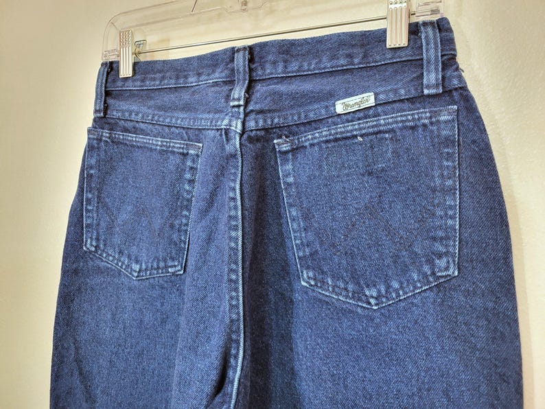 May include: Close-up of the back of dark blue denim jeans. The jeans have two back pockets and a small rectangular tag with the brand name "Wrangler". The jeans are hanging on a clear plastic hanger.