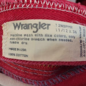 以下が含まれることがあります： 赤いWranglerの衣服のクローズアップ。縫い付けられたタグにはブランド名、お手入れ方法、および「Made in USA」が含まれています。衣服は綿100％で作られています。