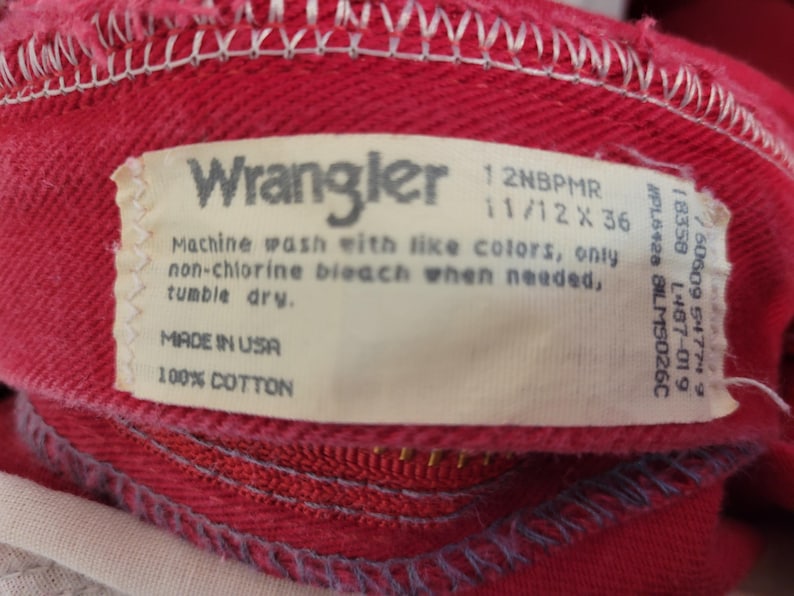 以下が含まれることがあります： 赤いWranglerの衣服のクローズアップ。縫い付けられたタグにはブランド名、お手入れ方法、および「Made in USA」が含まれています。衣服は綿100％で作られています。