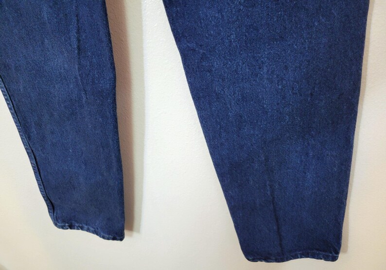 May include: Close-up of the lower portion of a pair of dark blue denim jeans. The jeans are slightly faded and have a classic, straight-leg fit. The fabric appears to be a medium-weight denim, suitable for everyday wear. The image shows the legs of the jeans.