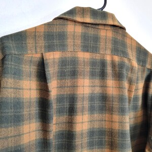 Pode incluir: Um close-up de uma camisa xadrez em tons de verde e laranja. A camisa est&aacute; pendurada em um cabide preto. O padr&atilde;o xadrez &eacute; um design cl&aacute;ssico, com gola e fechamento por bot&otilde;es.