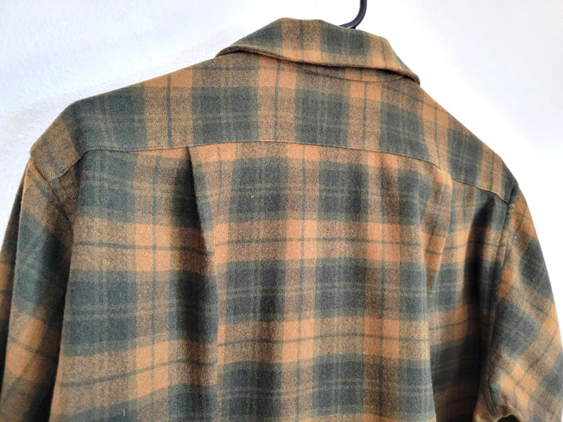 Pode incluir: Um close-up de uma camisa xadrez em tons de verde e laranja. A camisa est&aacute; pendurada em um cabide preto. O padr&atilde;o xadrez &eacute; um design cl&aacute;ssico, com gola e fechamento por bot&otilde;es.