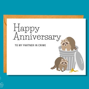 May include: A white card with the text "Happy Anniversary to my partner in crime" and a cartoon illustration of two raccoons in a trash can with a banana peel on the ground.