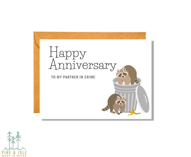 May include: A white card with the text "Happy Anniversary" and "To my partner in crime". The card features an illustration of two raccoons, one in a trash can and the other next to it.