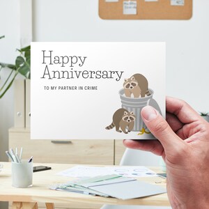 May include: A white card with the text "Happy Anniversary" and "To my partner in crime". Two cartoon raccoons are in a trash can with a banana peel on the ground.