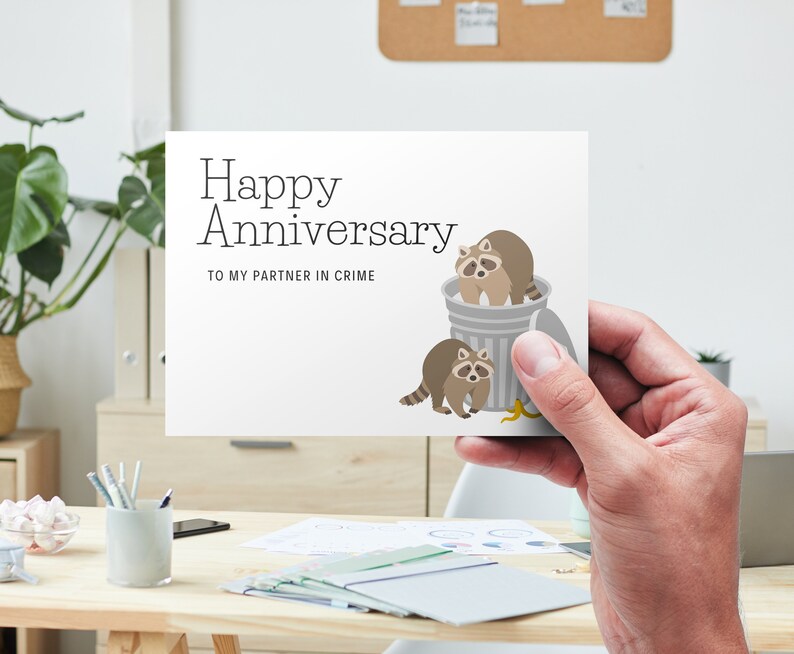 May include: A white card with the text "Happy Anniversary" and "To my partner in crime". Two cartoon raccoons are in a trash can with a banana peel on the ground.