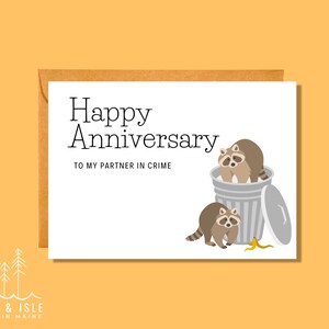 May include: A white card with the text "Happy Anniversary" and "To my partner in crime". The card features an illustration of two raccoons in a trash can.