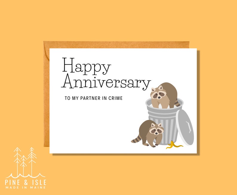 May include: A white card with the text "Happy Anniversary" and "To my partner in crime". The card features an illustration of two raccoons in a trash can.