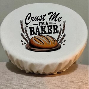 May include: White, circular fabric bread cover featuring the text "Crust Me I'm a BAKER" in black script. The design includes a loaf of bread on a wooden board, with wheat stalks. A great gift for bakers.