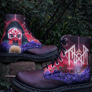 Può includere: Paio di stivali in pelle bordeaux con intricati disegni dipinti. Uno stivale presenta una figura incappucciata e accenti floreali, l'altro un design geometrico. Entrambi hanno lacci neri e dettagli di rose viola.