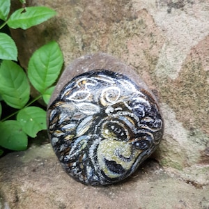 May include: Hand-painted rock art depicting a detailed portrait of a woman's face with a crown of leaves and intricate details. The rock is painted in shades of black, white, gold, and green.