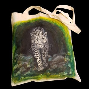 Könnte beinhalten: Eine weiße Stofftasche mit einem handgemalten Bild eines Leoparden, der aus einer dunklen Höhle kommt. Der Hintergrund ist grün und gelb.