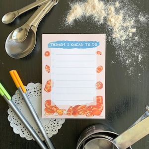 May include: A pink notepad with the text "Things I Knead To Do" and a checklist. The notepad is decorated with illustrations of various breads and pastries.