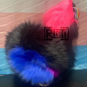 May include: A furry accessory with a black, blue, and hot pink color scheme. The accessory has a logo that says "Riot!". The background has a transgender pride flag.