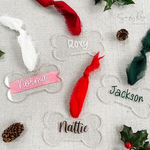 May include: Four clear acrylic dog bone ornaments with the names Roxy, Norma, Jackson, and Nattie written on them. Each ornament is decorated with a different colored ribbon.