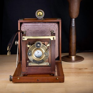 May include: A vintage wooden camera with a brass lens and a brass shutter speed dial. The camera is on a wooden table next to a copper lamp with a wooden base.