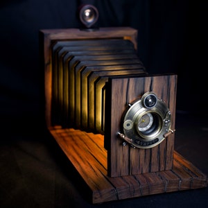 May include: A wooden camera with a bellows design and a brass lens. The camera is lit from within, casting a warm glow.