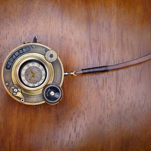 Puede incluir: Una cámara vintage de latón con una correa de cuero. La cámara tiene una cara redonda con un dial de reloj y las palabras "Compur" impresas en la cara.