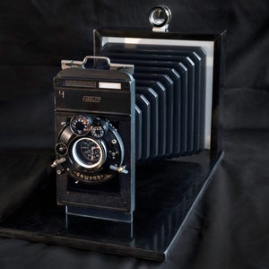 May include: A black vintage camera with a bellows system and a lens with multiple dials and gauges. The camera is on a black stand.