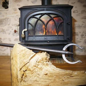 Peut inclure: Un tisonnier de cheminée en métal avec une extrémité incurvée et une longue poignée repose sur un morceau de bois. Un foyer noir avec un feu brûlant est en arrière-plan. La pièce a un mur en pierre.