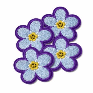 Peut inclure: Quatre écussons brodés en forme de fleurs. Chaque écusson présente un motif de pétales bleu clair avec une bordure violette et un centre jaune. Les écussons sont disposés sur un fond blanc.