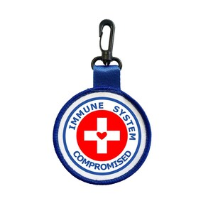 Peut inclure: Un écusson rond avec l'inscription "IMMUNE SYSTEM COMPROMISED" entourant une croix rouge avec un cœur au centre. L'écusson est blanc avec un bord bleu et une sangle bleue attachée à un clip noir.