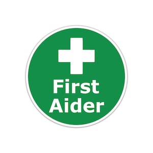 May include: A circular green sign featuring a white cross and the words "First Aider" in white. The sign has a white border and is set against a white background.