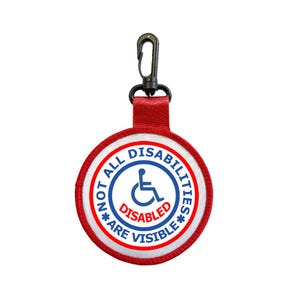 May include: A circular red, white, and blue fabric keychain with a black plastic lobster clasp. The center features a blue wheelchair symbol and the text NOT ALL DISABILITIES ARE VISIBLE in blue, with the word DISABLED in red at the center.