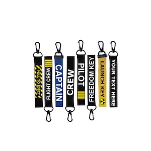May include: Set of seven black keychains with different coloured straps and text. The straps have the following text: "Pull to Eject", "Flight Crew", "Captain", "Crew", "Pilot", "Freedom Key", and "Launch Key". One strap is yellow with a black radiation symbol. One strap is blue. The rest are black.