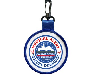 Puede incluir: Un parche de alerta médica redondo con el texto "MEDICAL ALERT", "SEIZURE DISORDER" y "FAINTING DISORDER". Tiene un borde azul y un clip negro.