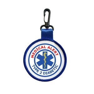 Puede incluir: Un parche de alerta médica redondo con un diseño azul y blanco. El parche tiene las palabras "MEDICAL ALERT" y "TYPE 2 DIABETIC" en un patrón circular. Un símbolo azul de la Estrella de la Vida está en el centro, con un bastón dorado. Tiene una correa azul y un clip negro.