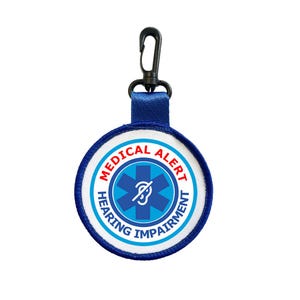 Puede incluir: Una etiqueta redonda de alerta médica con diseño azul y blanco. Contiene las palabras "MEDICAL ALERT" y "HEARING IMPAIRMENT". Incluye una correa azul y un clip negro.