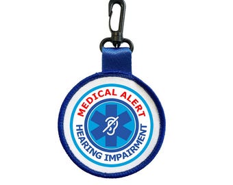 Puede incluir: Una etiqueta redonda de alerta médica con un diseño azul y blanco, con las palabras "MEDICAL ALERT" y "HEARING IMPAIRMENT". La etiqueta tiene una correa azul y un clip negro.