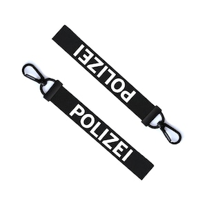 Pode incluir: Dois chaveiros pretos com fechos de metal preto. Cada chaveiro tem a palavra "POLIZEI" impressa em branco.
