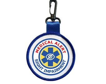 Puede incluir: Una etiqueta redonda de alerta médica con el texto "MEDICAL ALERT SIGHT IMPAIRMENT" en azul y rojo. La etiqueta presenta un símbolo azul de la estrella de la vida con un ojo en el centro. Tiene un lazo de tela azul y un clip negro.
