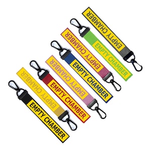 May include: A collection of fabric keychains in various colors, including black, yellow, red, pink, blue, green, and gray. Each keychain has a black clasp and the words "EMPTY CHAMBER" in yellow lettering.
