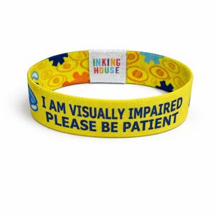 以下が含まれることがあります： 「I AM VISUALLY IMPAIRED PLEASE BE PATIENT」と紺色の文字が書かれた黄色の伸縮性リストバンド。青、オレンジ、黄色の図形が繰り返されるパターンです。「INKING HOUSE」のブランド名が白いラベルに印刷されています。