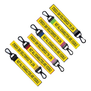 Op de afbeelding: Een set van zeven gele sleutelhangers met de tekst "NOT ALL DISABILITIES ARE VISIBLE" in zwart gedrukt. Elke sleutelhanger heeft een gekleurde streep.