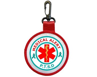 Puede incluir: Un parche redondo de alerta médica con el texto "MEDICAL ALERT" y "P.T.S.D.". El parche presenta una cruz roja con un símbolo de bastón y serpiente, rodeada por un borde turquesa y detalles de cinta. Tiene una correa roja y un clip negro.