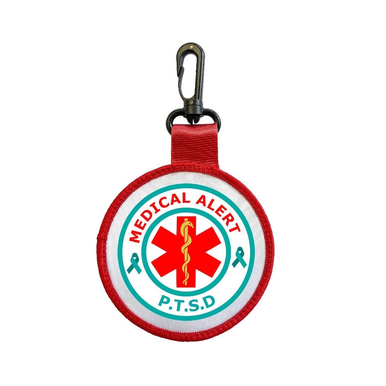 May include: A round medical alert patch with the text "MEDICAL ALERT" and "P.T.S.D." The patch features a red cross with a staff and snake symbol, surrounded by a teal border and ribbon accents. It has a red strap and a black clip.