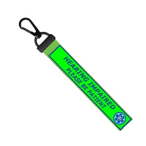 Può includere: Un cordino verde con la scritta "HEARING IMPAIRED PLEASE BE PATIENT" in bianco e blu. Il cordino ha una clip nera e un simbolo medico blu e bianco.
