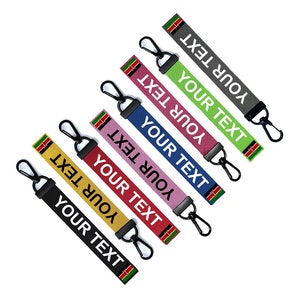 May include: Six colourful keychains with a black metal clasp and a white fabric strap with the text "YOUR TEXT" printed on it. Each keychain features a small Kenyan flag.